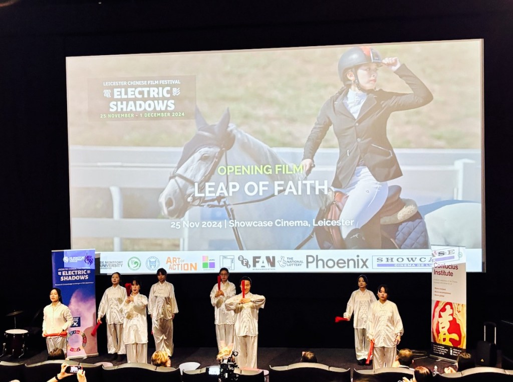 Tai Chi Fan Form performers on stage in front of cinema screen showing a rider sitting on a horse (still from the Chinese documentary “Leap of Faith”).